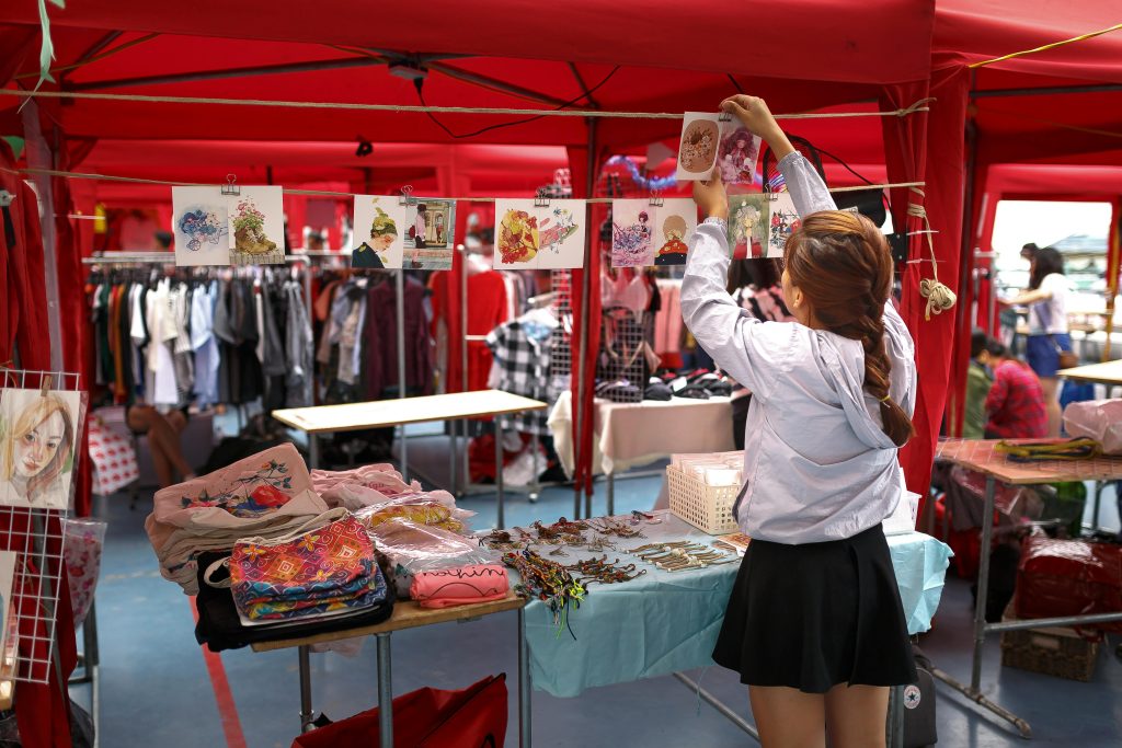 Home A woman organizes art prints in a vibrant outdoor market stall with colorful clothing and accessories.